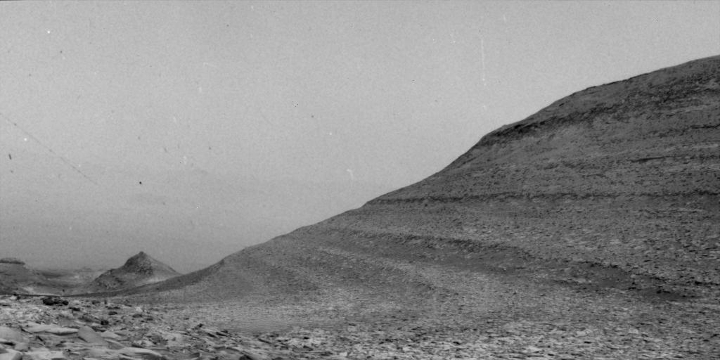 A black-and-white photo of a rocky Martian landscape, featuring a large sloped hill with visible layers on the right and a smaller peak in the distance under a hazy sky. Specks of white appear to dot the image from time to time, a response to solar energetic particles from the Sun hitting the camera.